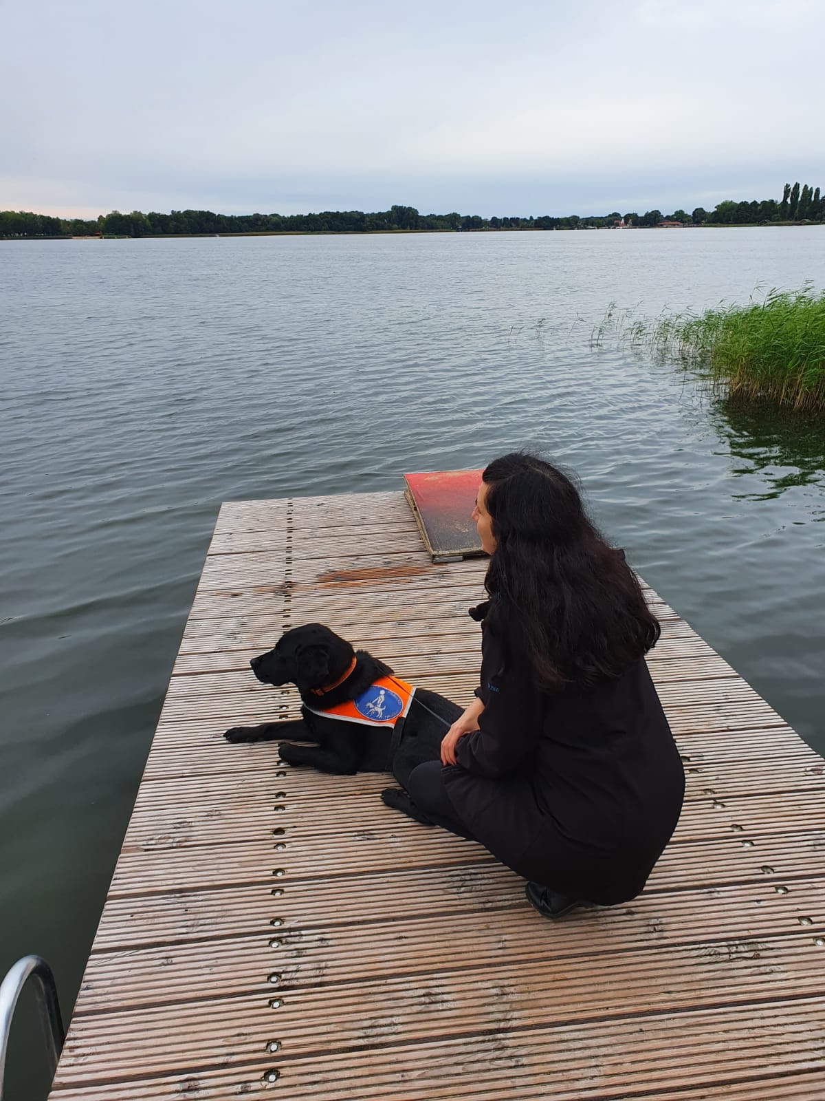 Jennifer Sonntag sitzt auf einem Steg am Wasser. Neben ihr liegt ihr Blindenführhund.