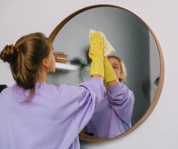 Eine Frau putzt einen runden spiegel. mit einem Lappen. Sie trägt Handschuhe und ist von Hinten und teilweise im Spiegel zu sehen.